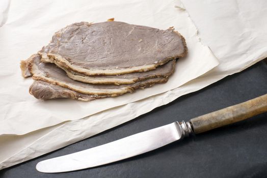 Pile of cold sliced roast beef for sandwiches on crumpled white paper with a kitchen knife in the foreground