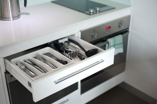 Open cutlery drawer in a kitchen cabinet with displayed kitchenware and eating utensils alongside a modern stove