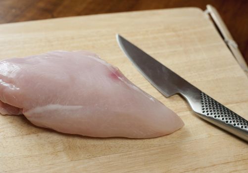 Uncooked lean healthy chicken breast on a wooden chopping board with a sharp kitchen knife , close up view