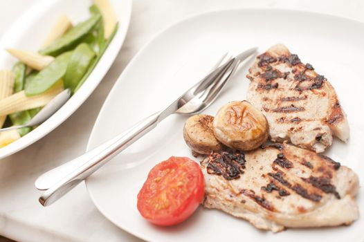 Grilled pork chops served with tomato and mushrooms and a side plate of and vegetables containging mangetout peas and baby corn