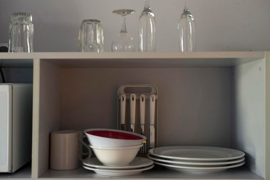 A variety of ceramic plates, mugs and bowls on a kitchen shelf together with some glasses stored above