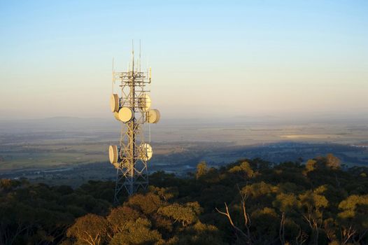 Mobile phone communications mast on a forested hilltop with dish antennae in an open landscape at sunset