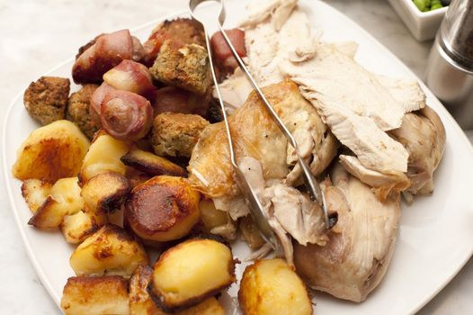 Carved roast chicken, roast potato and vegetables on a plate with metal thongs ready to be served for dinner