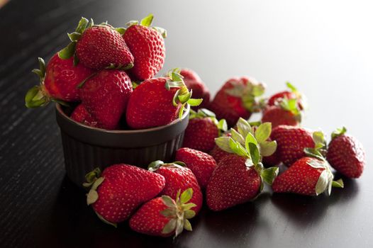 Fresh ripe red strawberries in a ramekin overflowing onto the surface below with side lighting, copyspace and shadow