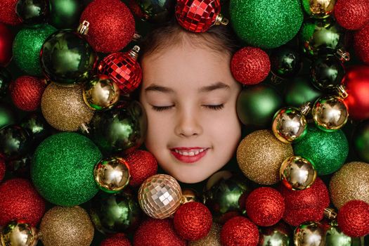 portrait of a little girl with closed eyes hidden in red, green and gold Christmas balls