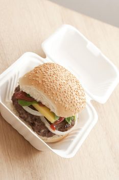 First person angled view on freshly prepared hamburger filled with onions, ketchup, cheese and lettuce in take out carton