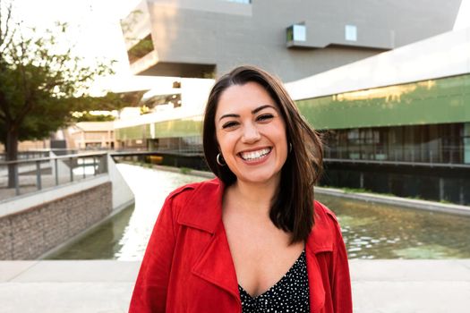 Portrait of smiling young brunette caucasian woman looking at camera wearing red jacket in a city park. Copy space. Headshot. Lifestyle concept.