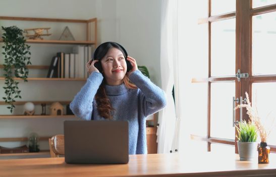 Happy Asian woman listening to music on computer while sitting in chair at home. smiling girl relaxing with headphones in the morning time to rest copy space.