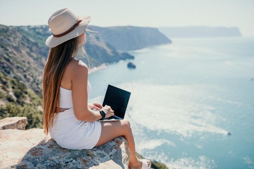 Successful business woman in yellow hat working on laptop by the sea. Pretty lady typing on computer at summer day outdoors. Freelance, travel and holidays concept.