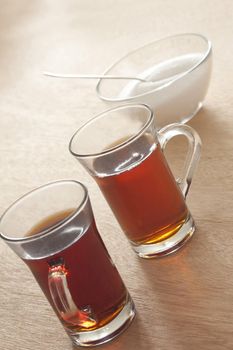 Two glass mugs of freshly brewed hot black tea with a bowl of sugar behind on a yellow background, tilted high angle view