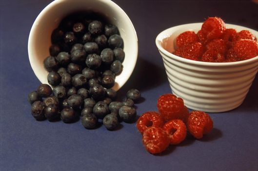 Delicious ripe fresh raspberries and blackcurrants in individual small dishes with the blackcurrants tilted on their side spilling onto the dark background