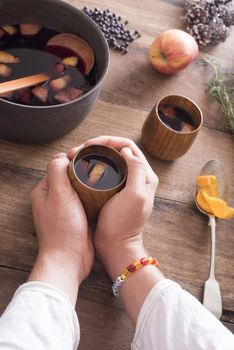 Person cradling a mug of homemade warm mulled wine for the holiday season with ingredients on a wooden table ad bowl of freshly made beverage alongside in a first person POV