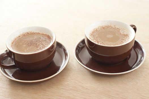 Two brown ceramic cups and saucers of fresh cappuccino coffee with a rich milky foam served on a wooden table with gradient color to white