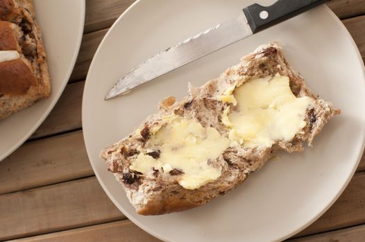 Buttered Hot Cross Bun for tea during the Easter festive season with an overhead view of a thickly buttered spicy fruity bun on a plate