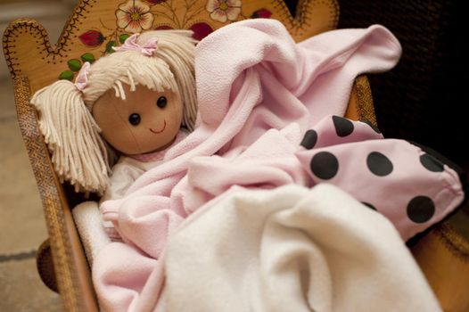 Blond toy doll wrapped in pink blankets lying in a wooden crib, close up view from above