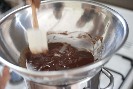 Melted chocolate in a stainless steel bowl being stirred by a white plastic baking spatula