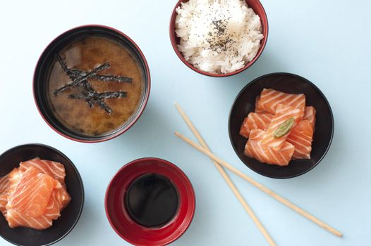 Overhead view of fresh cut and sliced salmon in small bowls beside soy dipping sauce and white rice by soup next to chopsticks against a light blue background