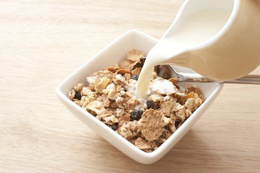 Jug of creamy white milk poured into breakfast cereal with raisins and nuts over light brown wooden table