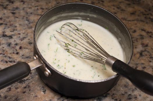 White creamy sauce seasoned with herbs in a saucepan with a metal hand whisk on a granite kitchen counter