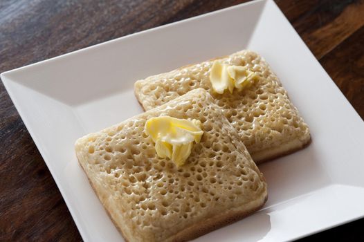 Two plain fried crumpets with butter on top neatly stacked partially over each other in simple matching square white plate