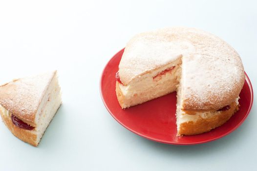 Fresh Victoria cream cake with strawberry jam served on a red plate with single slice cut and removed to the side over a white background