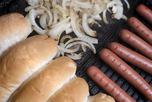 Cooking hot dog ingredients on a griddle with a row of smoked Wiener or Frankfurter sausages, diced onion and fresh white bread rolls