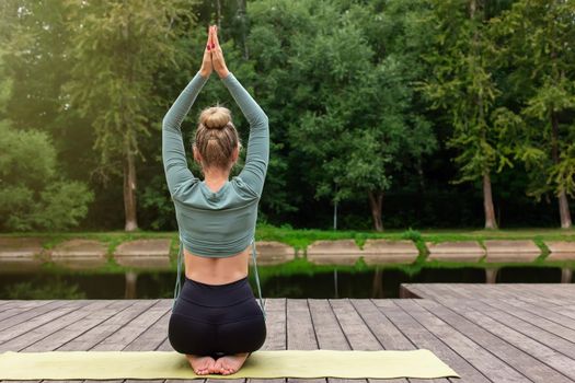 A some slender woman in a green top and black leggings, sitting on a sports mat, on a wooden platform in a park by a pond, with her hands raised up and her palms closed. Rear view. Copy space