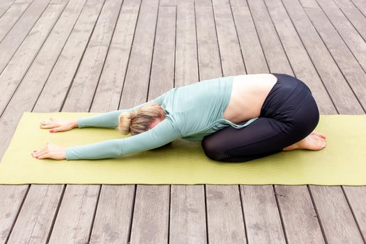 A blonde in a green top and leggings lies on a wooden platform by a pond in the park in summer and does yoga, performing the pose of a child.copy space. Top view