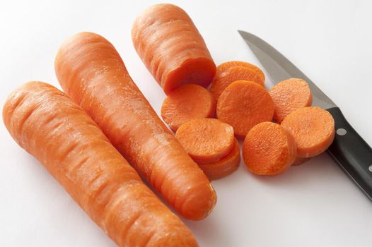 Preparing carrots for a meal slicing them with a sharp kitchen knife, close up high angle on three fresh carrots over white