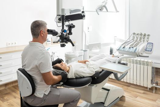 Male dentist using dental microscope treating patient teeth at dental clinic office. Medicine, dentistry and health care concept