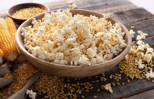 Wodden bowl of popcorn surrounded by popcorn kernels and popcorn on the cob