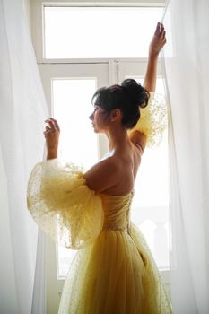 A woman's silhouette in a golden luxurious dress against the background of a window holds a curtain with her hands. Elegant lady in a yellow long silk dress with bare back, back view