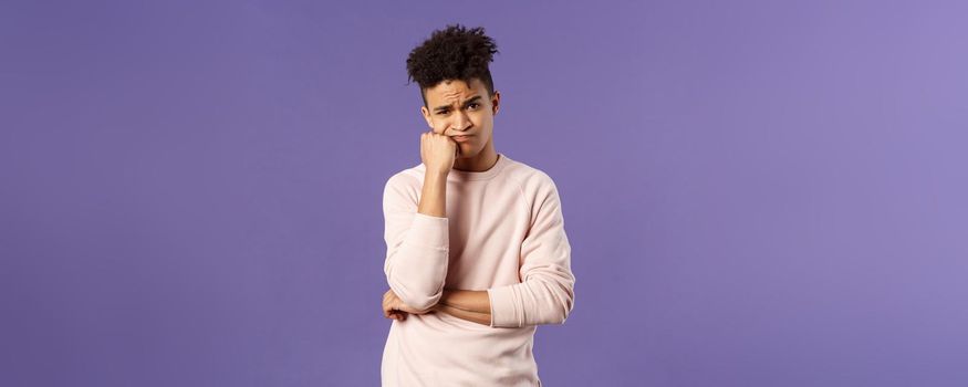 Portrait of complicated, troubled young hispanic man facing tough problem, lean on fist grimacing and pouting, solving troublesome situation, thinking, feeling uneasy, purple background.