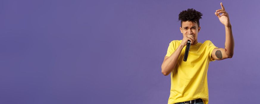 Portrait of young carefree sassy hipster guy with tattoos, yellow t-shirt performing in front audience with his hip-hop or rap, singing song, raising finger up, hold microphone, karaoke.