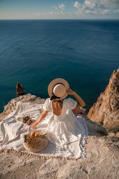 Street photo of a beautiful woman with dark hair in a white dress and hat having a picnic on a hill overlooking the sea.