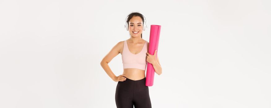 Sport, wellbeing and active lifestyle concept. Sassy beautiful asian fitness girl, woman ready for yoga classes or home training, holding rubber mat for workout and listening music in headphones.