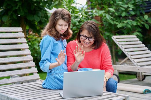 Mom and teenage daughter using laptop for chat conference video call. Parent and child relaxing together in backyard outdoor on lounger, laughing, waving hand at screen. Family technology lifestyle