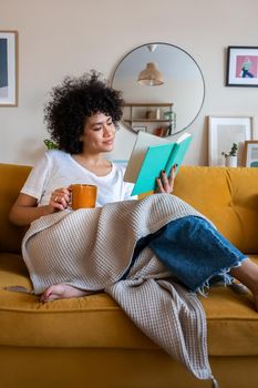 Relaxed, happy African american woman at home living room sitting on the couch, reading a book and drinking tea. Lifestyle concept.