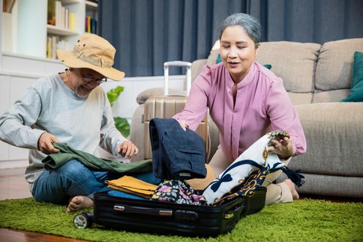 Asian romantic retired couple packing clothes travel bag suitcase together on floor at home interior living room, couple old senior married retired prepare luggage suitcase arranging for travel