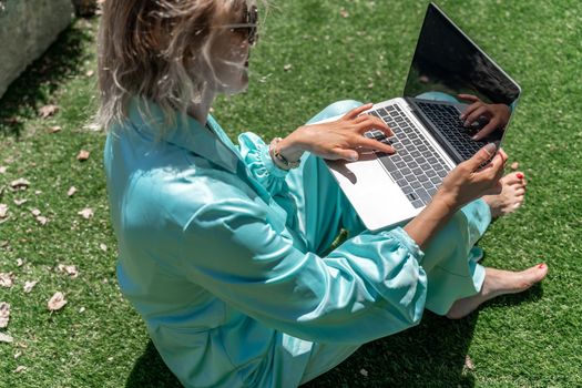 a young beautiful woman with blond curly hair in glasses and a blue dress sits on the grass in nature and uses a laptop