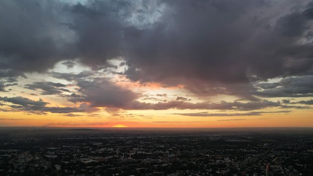 Large gray clouds in the orange light of the sun at sunset over the city of Almaty. A large city with low houses. Cars are driving on the road, lights are on. Green trees are growing. Drone view
