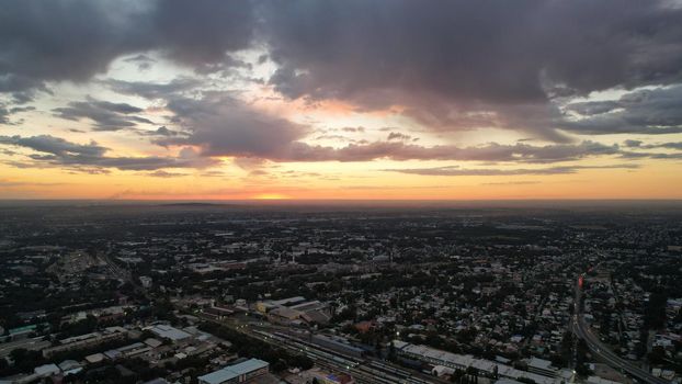 Large gray clouds in the orange light of the sun at sunset over the city of Almaty. A large city with low houses. Cars are driving on the road, lights are on. Green trees are growing. Drone view