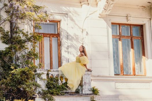 a beautiful smiling and kind woman in a gorgeous yellow dress stands on the balcony of an old vintage house.