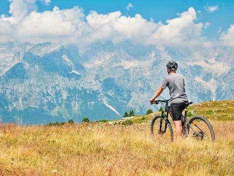 Teenager sit on modern mtb and watching from hill peak to opposite stonny and snowy peaks. Dolomite Alps, Trento region, Italy 
