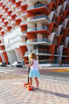 Dubai, UAE - 04.02.2021 - Little girl riding a scooter in front of Binghatti crystals residential building.