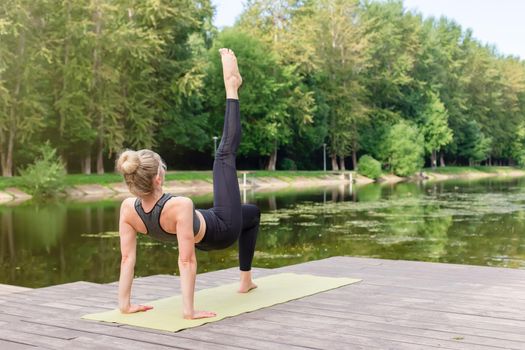 A woman in a gray top and leggings, on a wooden platform by a pond in a park in summer, does yoga, performing a table pose with her leg raised up. Copy space