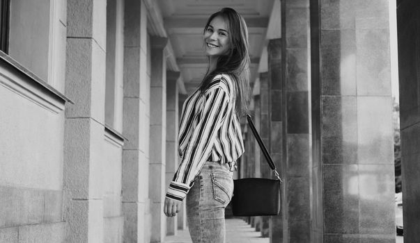 Street Style Outdoors Portrait of Beautiful Girl. Young Woman Smiling. Happy girl wearing Print Shirt, Jeans and Black Bag. Black and white shoot