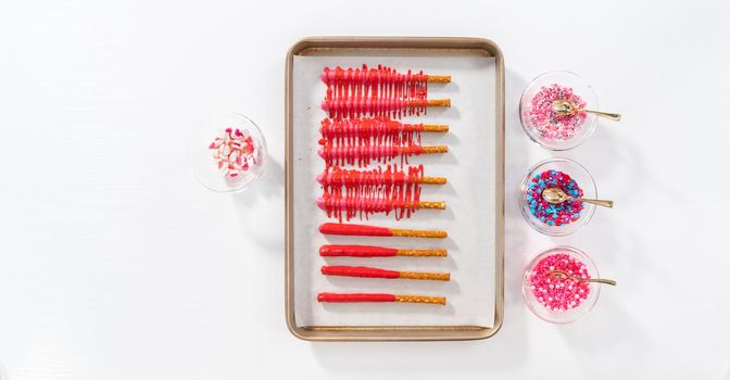 Flat lay. Drizzling melted chocolate over chocolate-dipped pretzels rods and decorating with sprinkles to make chocolate-covered pretzel rods for Valentine's Day.