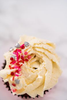 Piping white chocolate ganache frosting on top of red velvet cupcakes and topping with sprinkles.