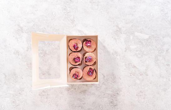 Flat lay. Packaging freshly baked chocolate strawberry cupcakes garnished with gourmet mini pink chocolates into a white paper cupcake box.